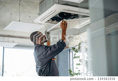 High up, fixing the ventilation. Repairman is indoors in the modern office 134381938