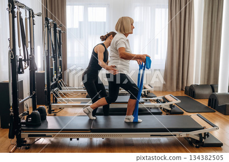 Blue colored resistance loops in hands for the legs. Female trainer is helping old woman to exercise 134382505