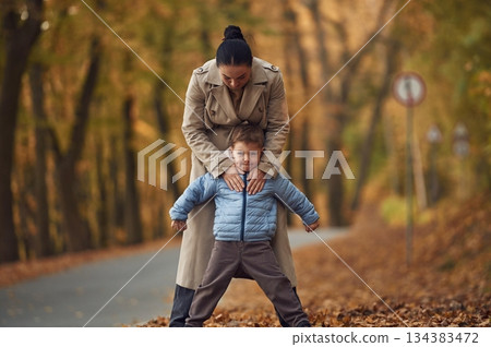 Fun times, behind the boy. Mother with her son are having a walk in autumn forest near the road Fun times, behind the boy. Mother with her son are having a walk in autumn forest near the road 134383472