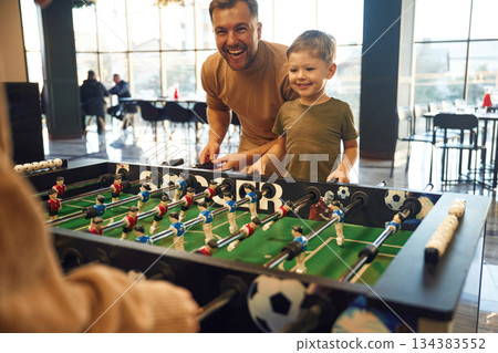 Smiling, enjoying. Family of dad, mother and son that are playing table football game indoors 134383552