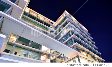Office building in the evening with lightened workspaces. Modern, glazed skyscraper in financial district. Late hours in the office. 134384428