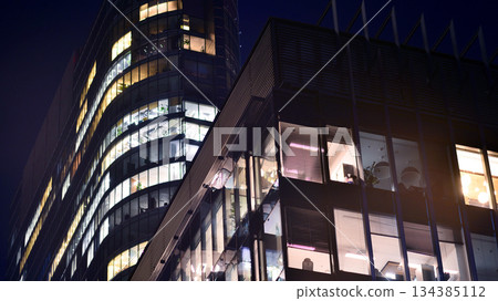Modern office building in city center illuminated at night. Rows of lit windows against the architectural grid of a modern facade. Concrete and glass facade in its night version. 134385112