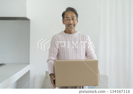 一位六十多歲的男子抱著一個紙箱,微笑地看著鏡頭。畫面展現了搬家和整理物品的過程。 一位六十多歲的男子抱著一個紙箱,微笑地看著鏡頭。畫面展現了搬家和整理物品的過程。 134387118