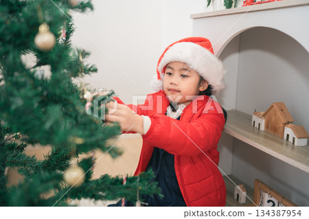 Little Girl in Red Jacket and Santa Hat Decorates Christmas Tree with Ornaments, Festive Atmosphere in Cozy Holiday Setting 134387954