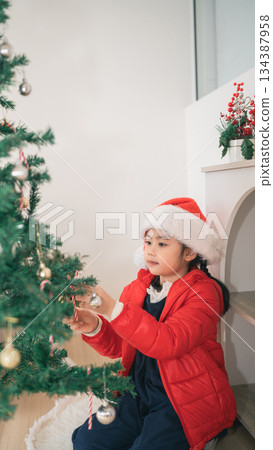 A Young Girl in a Christmas Hat Decorates a Festive Tree with Ornaments and Joyful Anticipation for the Holiday Season and Family Celebrations 134387958