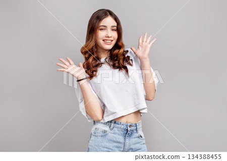 Beautiful teenage girl with wavy brown hair smiling and raising hands in playful gesture, posing in studio on gray background, young female fashion model in casual white top and denim 134388455