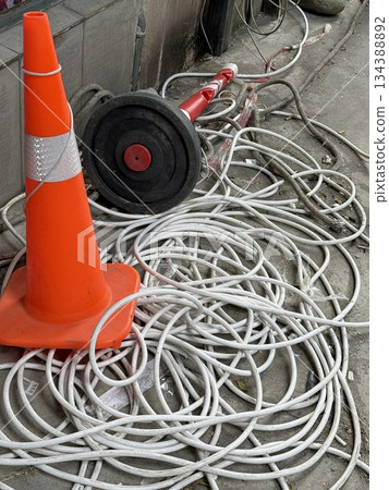 Traffic cone and tangled cables on concrete pavement, urban maintenance and safety concept Traffic cone and tangled cables on concrete pavement, urban maintenance and safety concept 134388892