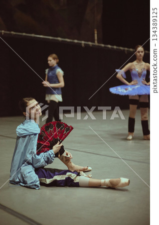 Ballet dancer sitting on floor holding decorative fan during break. Ballet dancer sitting on floor holding decorative fan during break. 134389125