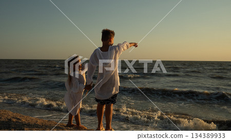 Two siblings stand together on a sandy beach at sunset, holding hands and pointing toward the open ocean. The children share a moment of admiration and connection with nature on the seashore. Two siblings stand together on a sandy beach at sunset, holding hands and pointing toward the open ocean. The children share a moment of admiration and connection with nature on the seashore. 134389933