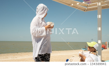Two children in white shirts eat snacks on a sunny sandy beach under an umbrella, admiring the calm ocean during a family summer vacation. The children snack on the beach between swims. 134389941
