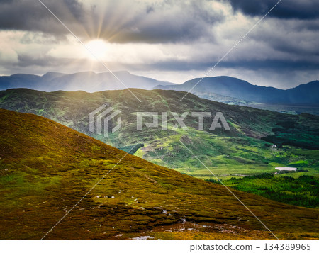 A scenic view of rolling hills and mountains against a dramatic sky. A bright ray of sunshine breaks through the dark clouds and illuminates the landscape of County Kerry. 134389965