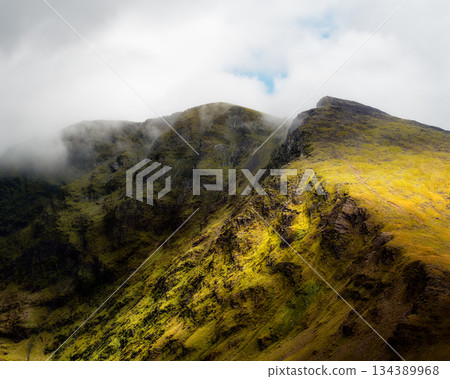 This scenic shot showcases the rugged terrain of the Coomloughra Horseshoe Mountains. Green grass covers the slopes and hiking trails are clearly visible. Clouds partially obscure the mountain peaks. 134389968