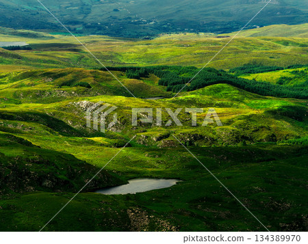 A wide angle showcases the lush, rolling green hills and small lake along the popular Coomloughra Horseshoe hiking trail, part of a vast scenic vista in Ireland. 134389970