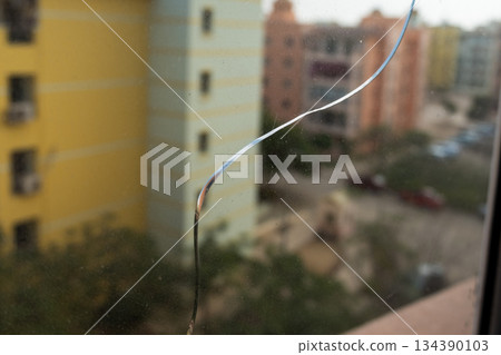 A close-up view of a crack on a glass window. The background shows blurred buildings under a clear blue sky. The glass surface reflects light. 134390103