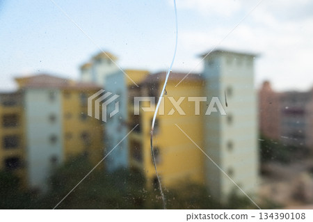A close-up view of a crack on a glass window. The background shows blurred buildings under a clear blue sky. The glass surface reflects light. A close-up view of a crack on a glass window. The background shows blurred buildings under a clear blue sky. The glass surface reflects light. 134390108