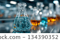 Two flasks on a lab bench filled with bubbling blue and amber liquids. Soft bokeh highlights a busy scientific workspace 134390352