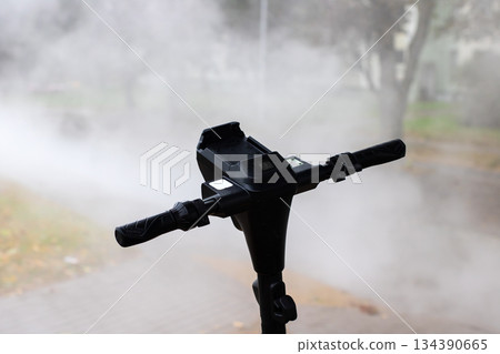Solitary scooter handlebar in foggy park with wet pavement and distant blurred green trees 134390665