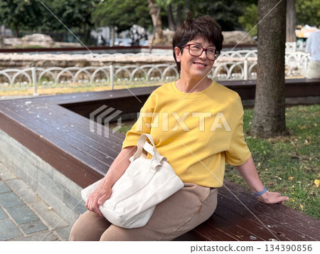 Smiling middle-aged woman resting on bench in green urban park in Istanbul. Positive mood, everyday happiness, active aging, and relaxed lifestyle in public outdoor environment. Smiling middle-aged woman resting on bench in green urban park in Istanbul. Positive mood, everyday happiness, active aging, and relaxed lifestyle in public outdoor environment. 134390856