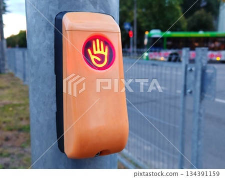 Pedestrian Traffic Light Push Button with Red Don't Walk Hand Signal. Close-up of orange pedestrian crossing button showing illuminated red hand "stop" signal on city street pole. Pedestrian Traffic Light Push Button with Red Don't Walk Hand Signal. Close-up of orange pedestrian crossing button showing illuminated red hand "stop" signal on city street pole. 134391159