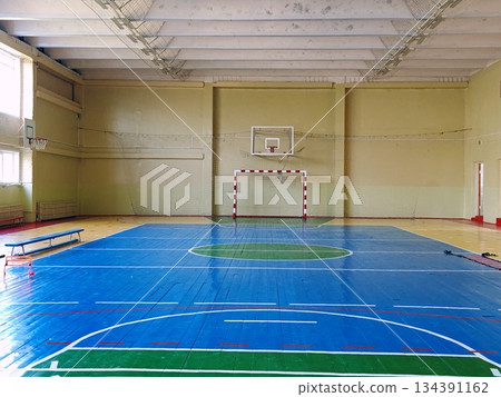 Empty School Gymnasium with Basketball and Handball Court Markings. Spacious empty indoor school sports hall with blue wooden floor, basketball hoops and handball goal. Concept of education, sport Empty School Gymnasium with Basketball and Handball Court Markings. Spacious empty indoor school sports hall with blue wooden floor, basketball hoops and handball goal. Concept of education, sport 134391162