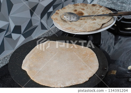 Making an unleavened flatbread the roti. A close-up of the round flatbread being fried on a flat, rimless griddle called a tawa, and several cooked roti on a white plate. Baking homemade bread. Making an unleavened flatbread the roti. A close-up of the round flatbread being fried on a flat, rimless griddle called a tawa, and several cooked roti on a white plate. Baking homemade bread. 134391204