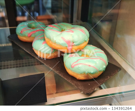 Colorful Glazed Donuts in Bakery Display. Stack of fresh turquoise donuts with pink icing drizzle displayed behind glass in a bakery or cafe, tempting sweet treat Colorful Glazed Donuts in Bakery Display. Stack of fresh turquoise donuts with pink icing drizzle displayed behind glass in a bakery or cafe, tempting sweet treat 134391264