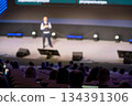 Speaker on stage presenting at conference as audience watches in a large auditorium 134391306