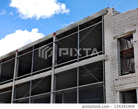 Unfinished Multi-Story Building Under Construction. Modern multi-floor concrete building under construction with exposed floors and brick walls against blue sky. 134391409