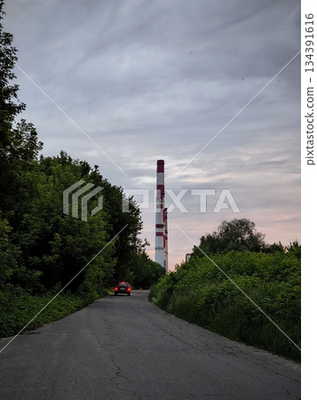 Industrial chimney with car on road. Tall red and white striped industrial chimney rising above green trees with a car driving on rural road under cloudy sky. Industrial chimney with car on road. Tall red and white striped industrial chimney rising above green trees with a car driving on rural road under cloudy sky. 134391616