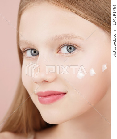 Portrait of a smiling cheerful girl applying face cream on cheek for skin glow. Advertising face cream and skincare products for teens. Blonde girl with blue eyes isolated in studio on pink background Portrait of a smiling cheerful girl applying face cream on cheek for skin glow. Advertising face cream and skincare products for teens. Blonde girl with blue eyes isolated in studio on pink background 134391764