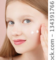 Portrait of a smiling cheerful girl applying face cream on cheek for skin glow. Advertising face cream and skincare products for teens. Blonde girl with blue eyes isolated in studio on pink background 134391766