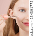 Half face portrait of a funny smiling young girl applying serum on face using an dropper. Blonde Caucasian girl with blue eyes isolated on pink background. Advertising concept skin care for teenagers. 134391772