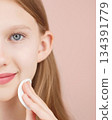 Half face portrait of smiling young girl using cotton pad in studio on pink background. Adorable Caucasian girl using cotton round cleansing skin. Concept of skincare advertising for teens. Copy space 134391779