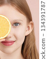 Half face portrait of a cheerful smiling blonde posing, studio holding an orange in front of her eyes on a pink background. Gen Alpha skincare ads. Haircare products for teenagers, children. Selfcare 134391787