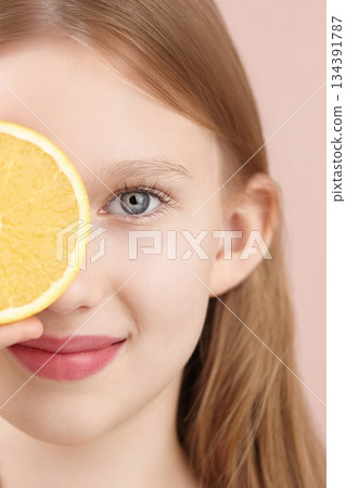 Half face portrait of a cheerful smiling blonde posing, studio holding an orange in front of her eyes on a pink background. Gen Alpha skincare ads. Haircare products for teenagers, children. Selfcare Half face portrait of a cheerful smiling blonde posing, studio holding an orange in front of her eyes on a pink background. Gen Alpha skincare ads. Haircare products for teenagers, children. Selfcare 134391787