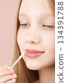 Cute young girl applying lip gloss. Caucasian blonde girl gen Alpha with blue eyes using make-up cosmetics, posing in studio on pink background. Lipstick. Concept of advertising cosmetics for teens 134391788