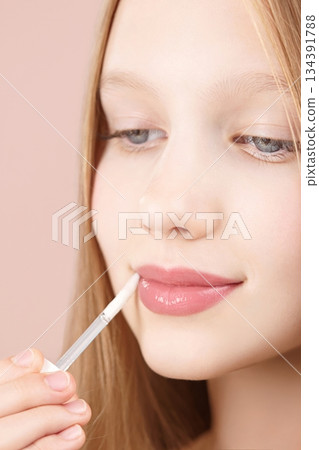 Cute young girl applying lip gloss. Caucasian blonde girl gen Alpha with blue eyes using make-up cosmetics, posing in studio on pink background. Lipstick. Concept of advertising cosmetics for teens 134391788