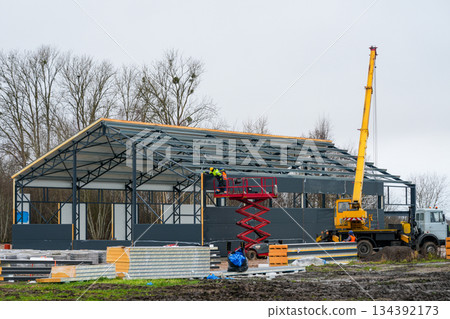 Construction workers install wall panels on a steel industrial building frame using lift and crane 134392173