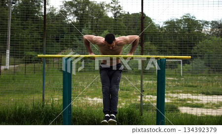Hardy shirtless sportsman doing exercises on parallel bars at nature. Strong and muscular man working out at sports ground. Young athletic guy exercising outdoor. Concept of active healthy lifestyle 134392379