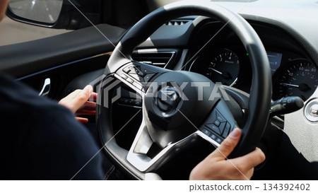 Male arms holds steering wheel while driving a motor car at city. Young man operating the automobile at urban road. Driver moving in his auto on route. Slow motion 134392402