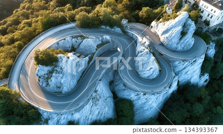 Winding Asphalt Road Curves Through Mountains With Lush Greenery Surrounding Rocky Cliffs Winding Asphalt Road Curves Through Mountains With Lush Greenery Surrounding Rocky Cliffs 134393367