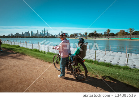 Cycling By Water, Parent And Child Cycling Along Lakeside With City Views And Relaxed Vibe 134393539