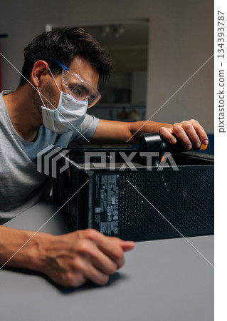 Vertical portrait of computer technician in protective mask and safety eyewear cleaning system unit with vacuum during precision hardware maintenance. Servicing motherboard of dusty computer. Vertical portrait of computer technician in protective mask and safety eyewear cleaning system unit with vacuum during precision hardware maintenance. Servicing motherboard of dusty computer. 134393787