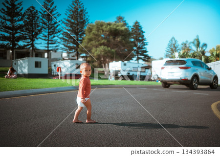 young child confidently strolling through sunny caravan park surrounded by tall pine trees 134393864