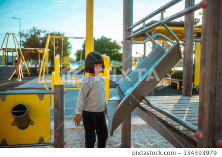 child stands thoughtfully on playground slide with warm sunset glow illuminating scene 134393979