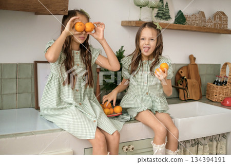 Children play with tangerines, laughing mischievously in the cozy kitchen, decorated for Christmas and New year. Children play with tangerines, laughing mischievously in the cozy kitchen, decorated for Christmas and New year. 134394191