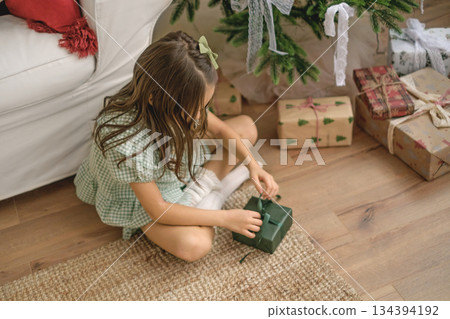 A child unties the green bow of a gift wrapped in holiday paper while sitting on the floor by the Christmas tree. 134394192