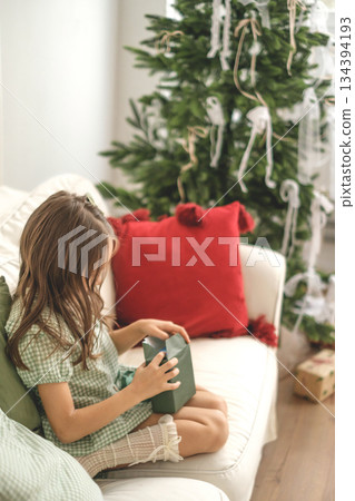 A child unwraps a paper wrapper of a Christmas gift in the early morning, sitting on the couch by a fir tree A child unwraps a paper wrapper of a Christmas gift in the early morning, sitting on the couch by a fir tree 134394193