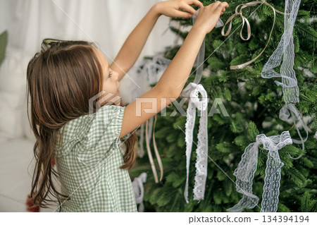 A girl ties a lace ribbon bow on a branch of a Christmas tree. Preparing for the New Year 134394194