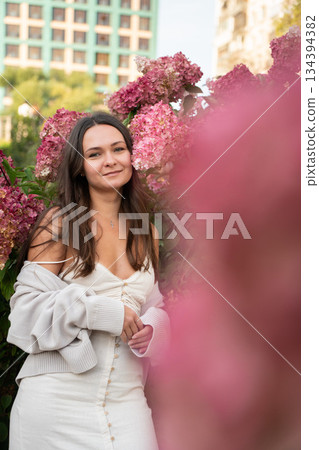 Young woman in a light dress stands among pink hydrangea flowers in an urban garden, soft daylight, pastel tones, calm and warm mood 134394382